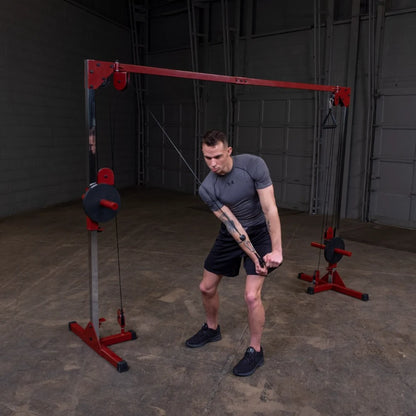 A man in a gray athletic shirt and black shorts performs a cable exercise using a red and silver pulley system in a gym setting.