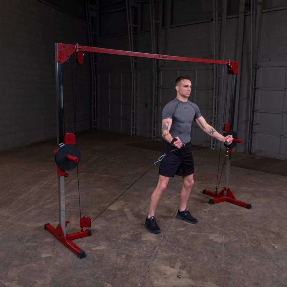 A young man with tattoos wearing a gray athletic shirt and black shorts is performing a cable exercise in a gym, using a cable machine with red and silver components. The background features a spacious, empty gym area.