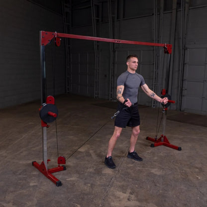 A young man with tattoos wearing a gray athletic shirt and black shorts is performing a cable exercise in a gym, using a cable machine with red and silver components. The background features a spacious, empty gym area.