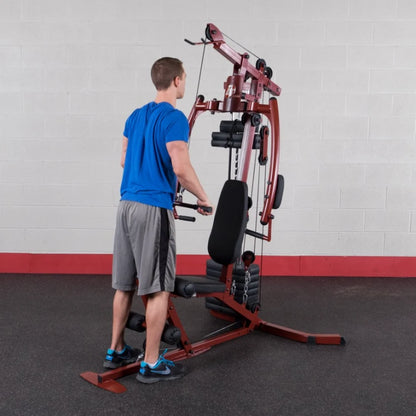 A man in a blue shirt and gray shorts is exercising on a multi-gym machine in a gym setting, focusing on strength training.