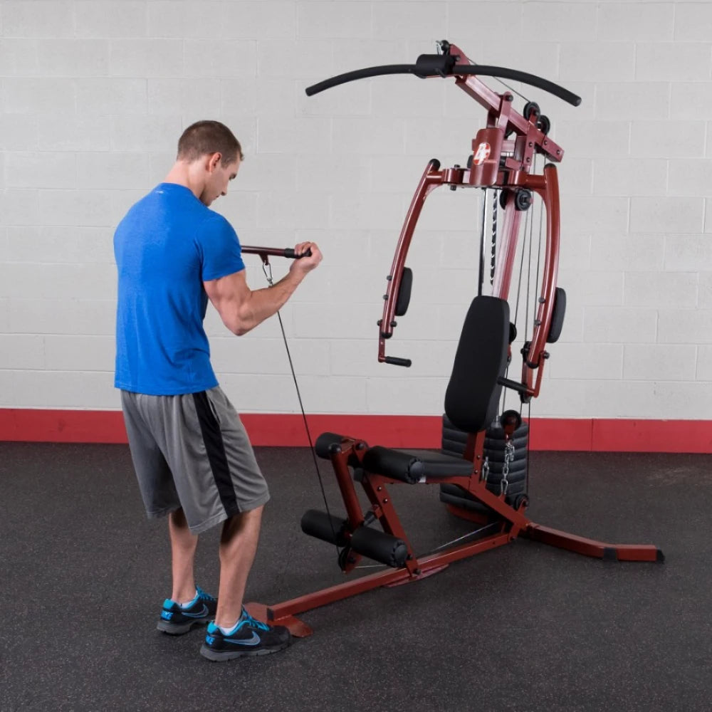 A man in a blue shirt and gray shorts is exercising on a multi-gym machine, performing a cable row. The gym has a simple design with a gray wall and red accents.
