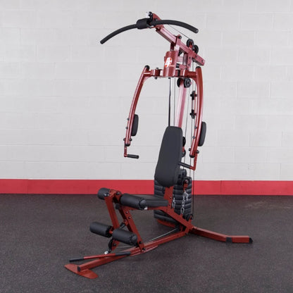 A multi-functional home gym equipment in a red and black color scheme, featuring a pull-up bar, adjustable seat, and various pulleys for strength training, set against a plain wall with a red base.