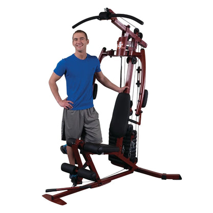 A young man in a blue shirt and gray shorts stands next to a multi-station home gym equipment, smiling confidently.