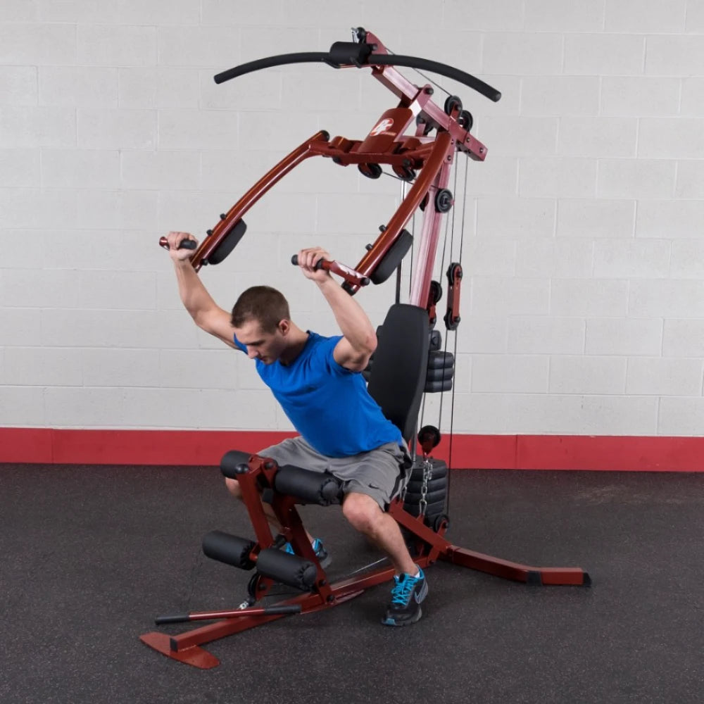 A man in a blue shirt using a multi-gym exercise machine, performing a seated workout with his arms extended upward, in a gym setting with gray walls and red accents.