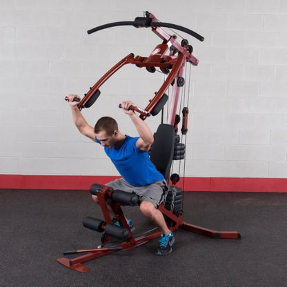 A man in a blue shirt using a multi-gym exercise machine, performing a seated workout with his arms extended upward, in a gym setting with gray walls and red accents.