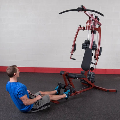A man in a blue shirt and gray shorts is seated on a gym machine, performing a row exercise. The machine features multiple pulleys and weights, set against a plain wall with a red base.