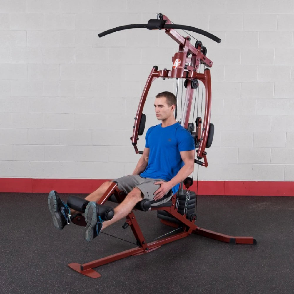 A man in a blue shirt and gray shorts using a multi-gym machine for leg exercises in a fitness facility with a gray wall and red accents.