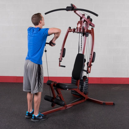 A person in a blue shirt and gray shorts using a multi-gym exercise machine in a gym setting, performing a workout that involves pulling a cable attachment. The background features a plain wall with a red base.