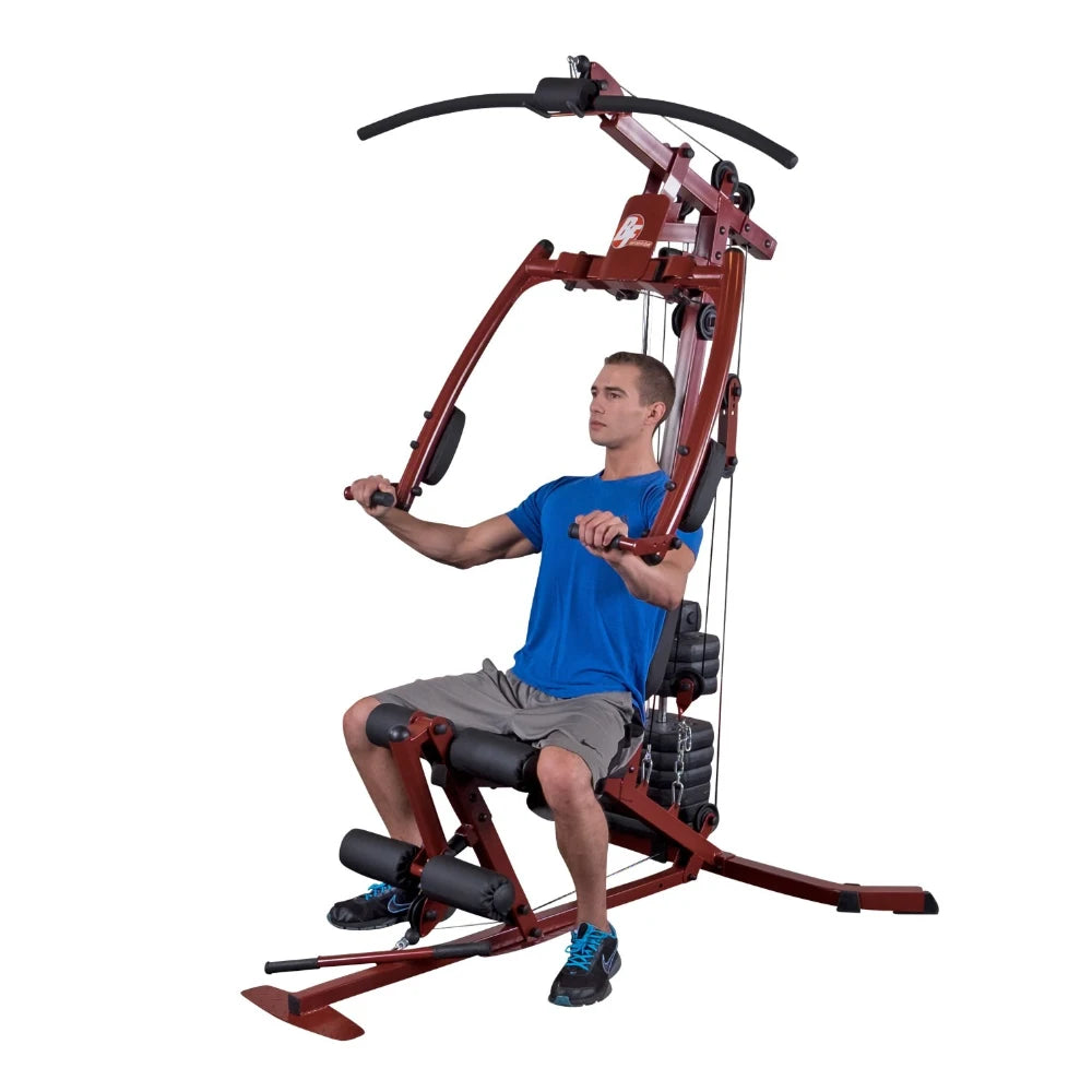 A young man in a blue shirt and gray shorts is using a multi-gym exercise machine, performing a seated workout with adjustable cables and weights.