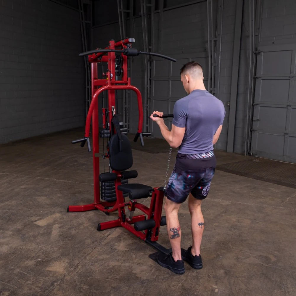 A person in a fitted gray shirt and patterned shorts performs a bicep curl using a red multi-gym machine in a spacious gym environment.