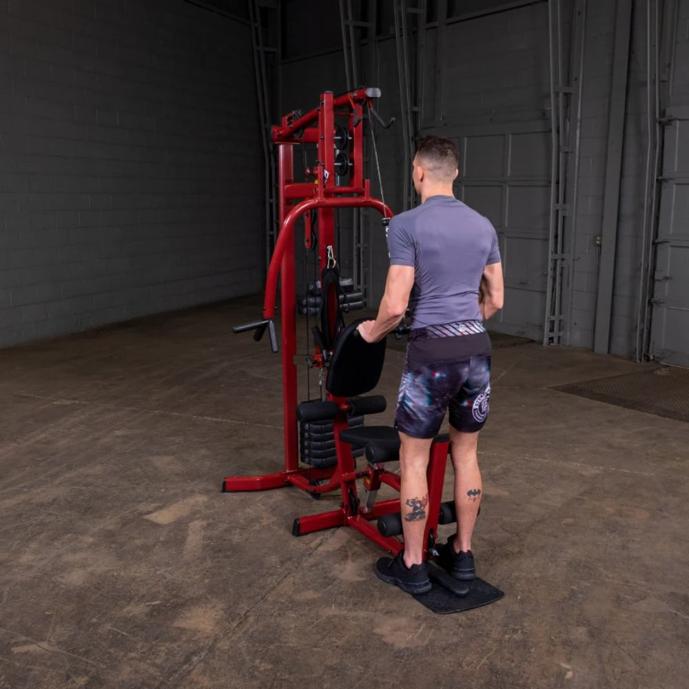 A person standing in front of a red multi-gym machine in an indoor gym setting, wearing a gray athletic shirt and patterned shorts.