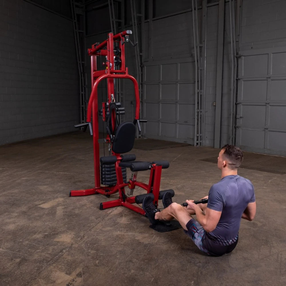 A person exercising on a red multi-gym machine, sitting on the floor and holding a handle, in an industrial-style gym environment.