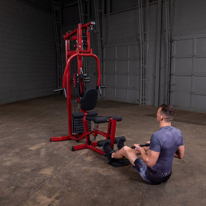 A person exercising on a red multi-gym machine, sitting on the floor and holding a handle, in an industrial-style gym environment.