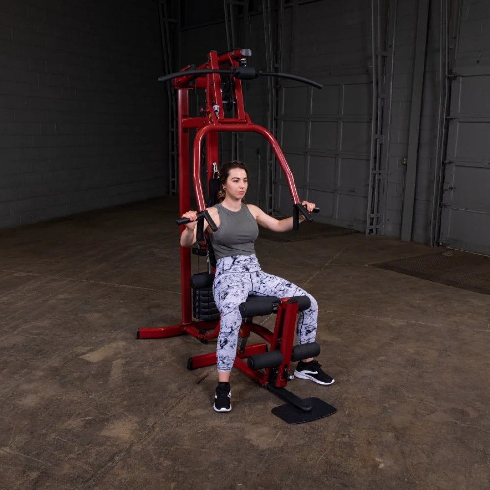 A woman performing an exercise on a red gym machine, wearing a gray tank top and patterned leggings, in a spacious indoor gym setting.