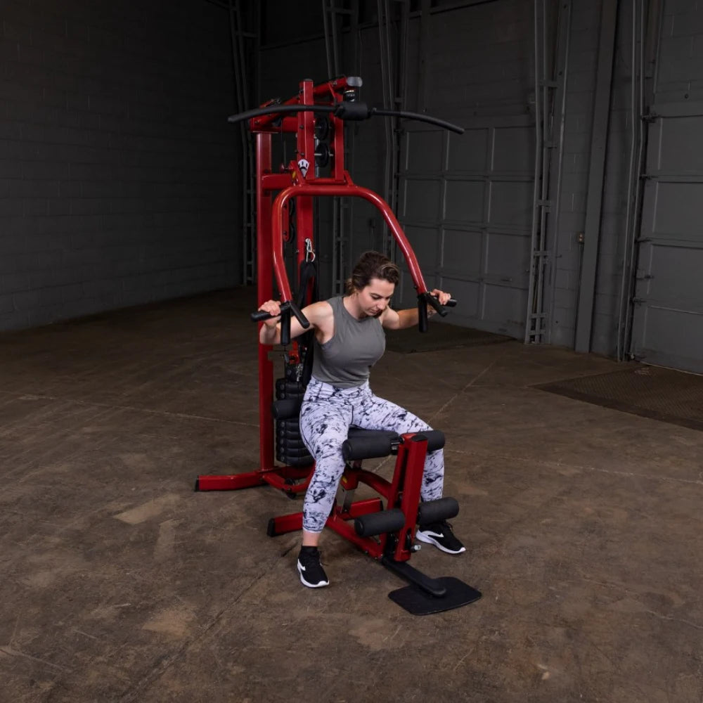 A woman in athletic wear is using a red weight machine in a gym, focusing on her workout. She is seated, with her hands gripping the machine's handles, in a spacious indoor area with a concrete floor.