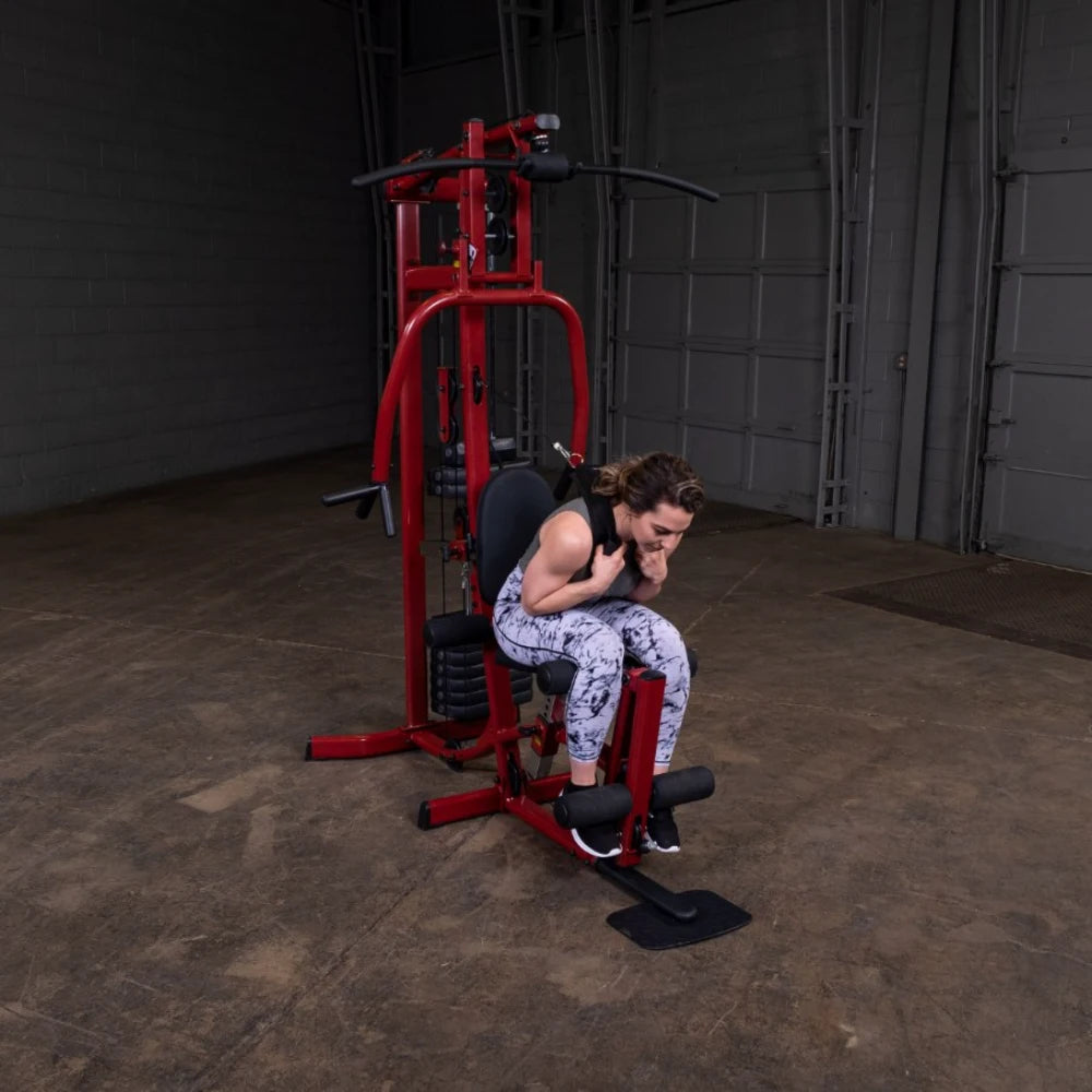 A woman in athletic wear performing an exercise on a red multi-gym machine in a spacious indoor gym setting.