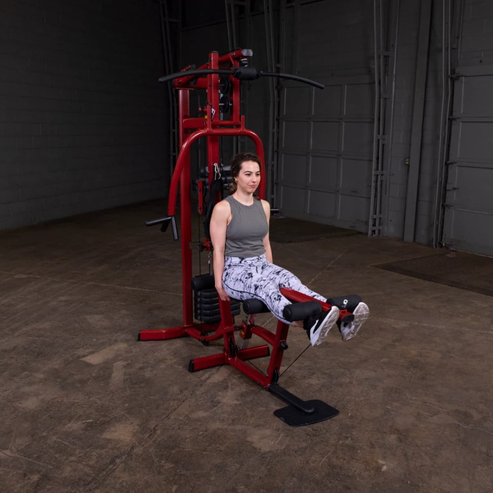 A woman in workout attire is seated on a red multi-gym machine, preparing to perform an exercise. The gym is set in a spacious, industrial-style environment with gray walls and a concrete floor.