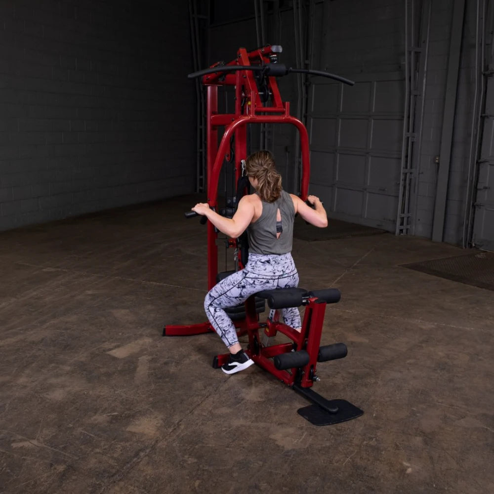 A person exercising on a red gym machine in a spacious indoor setting, wearing a gray tank top and patterned leggings, with a focus on their back and arms as they pull the machine's handles.