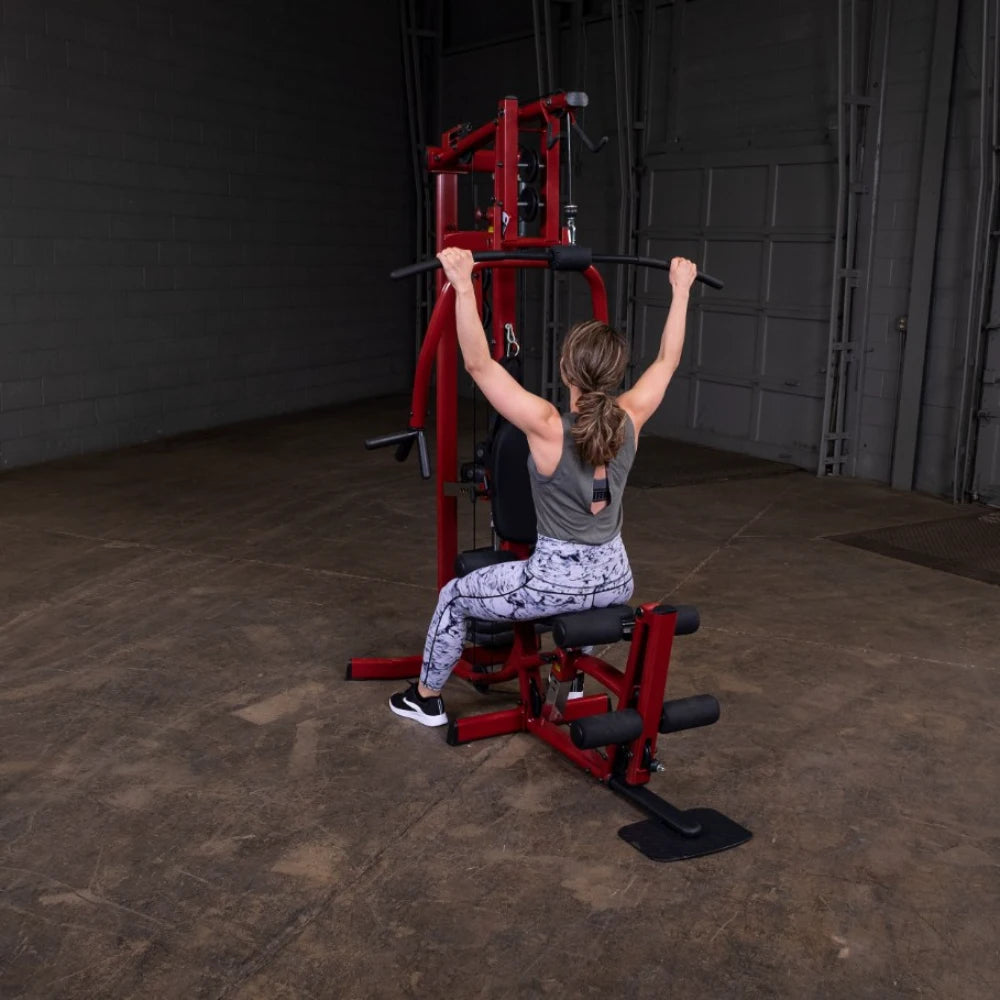 A woman performing an exercise on a multi-gym machine, seated with her back to the camera, pulling down the bar with both hands. She is wearing a gray tank top and patterned leggings, in a spacious gym environment with a concrete floor and gray walls.