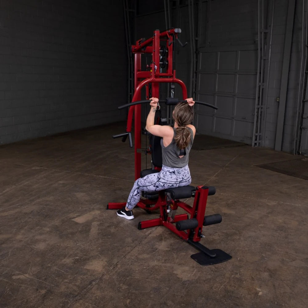A woman exercising on a red multi-gym machine, performing a pull-down exercise, wearing a gray tank top and patterned leggings, in a spacious gym environment.