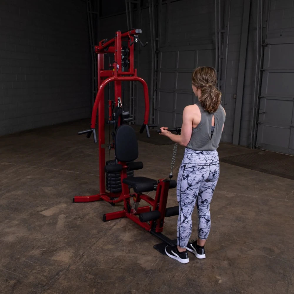 A woman with long hair tied in a ponytail is using a cable machine in a gym setting. She is wearing a gray tank top and patterned leggings, facing away from the camera while pulling a cable attachment towards her. The gym has a spacious, industrial look with concrete flooring and a large, open area.