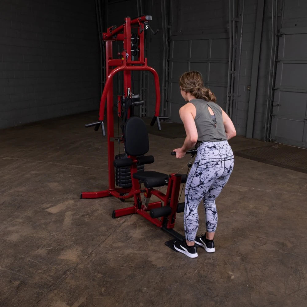 A person in athletic wear is using a red multi-gym machine in a spacious gym. The individual is positioned to perform a seated exercise, focusing on strength training. The background features a gray wall and a concrete floor.