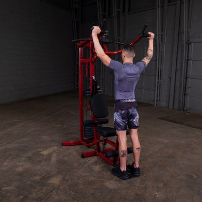 A person performing a lat pull-down exercise on a red gym machine, wearing a fitted gray shirt and patterned shorts, in a spacious indoor gym setting.