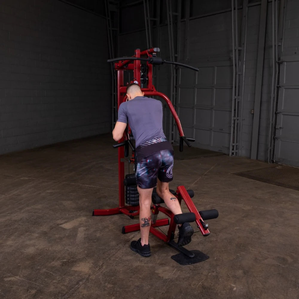 A person wearing a gray shirt and patterned shorts using a red exercise machine in a spacious gym setting.