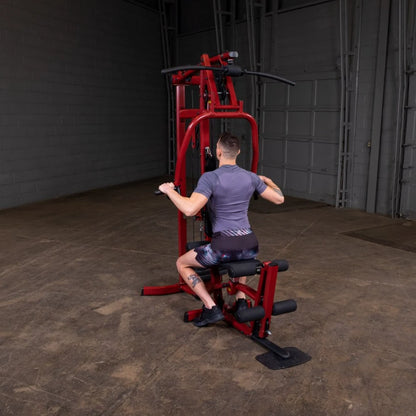 A person performing an exercise on a red gym machine, seated with hands gripping the handles, in an industrial-style gym environment.