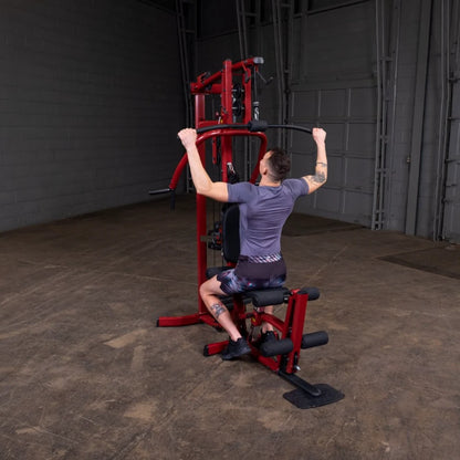 Person performing a lat pull-down exercise on a red gym machine in an industrial-style gym setting.