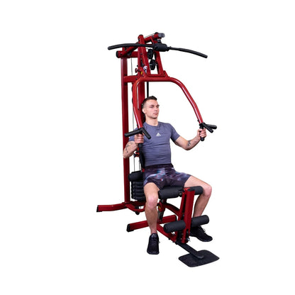 Man exercising on a red multi-gym machine, performing a chest press while seated, wearing a gray shirt and patterned shorts.