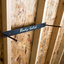 Black metal wall hanger with "Body-Solid" text mounted on OSB board, designed to hold foam mats vertically.