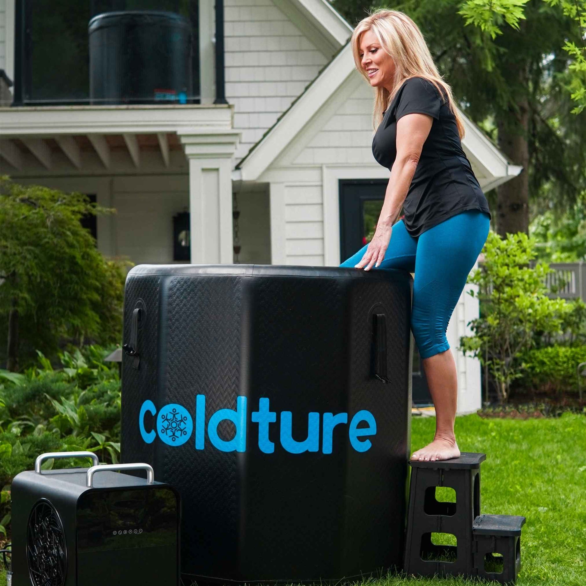 A woman in athletic wear stands on a stool beside a Coldture Barrel + Chiller Pro, set in a lush green yard with a house in the background.