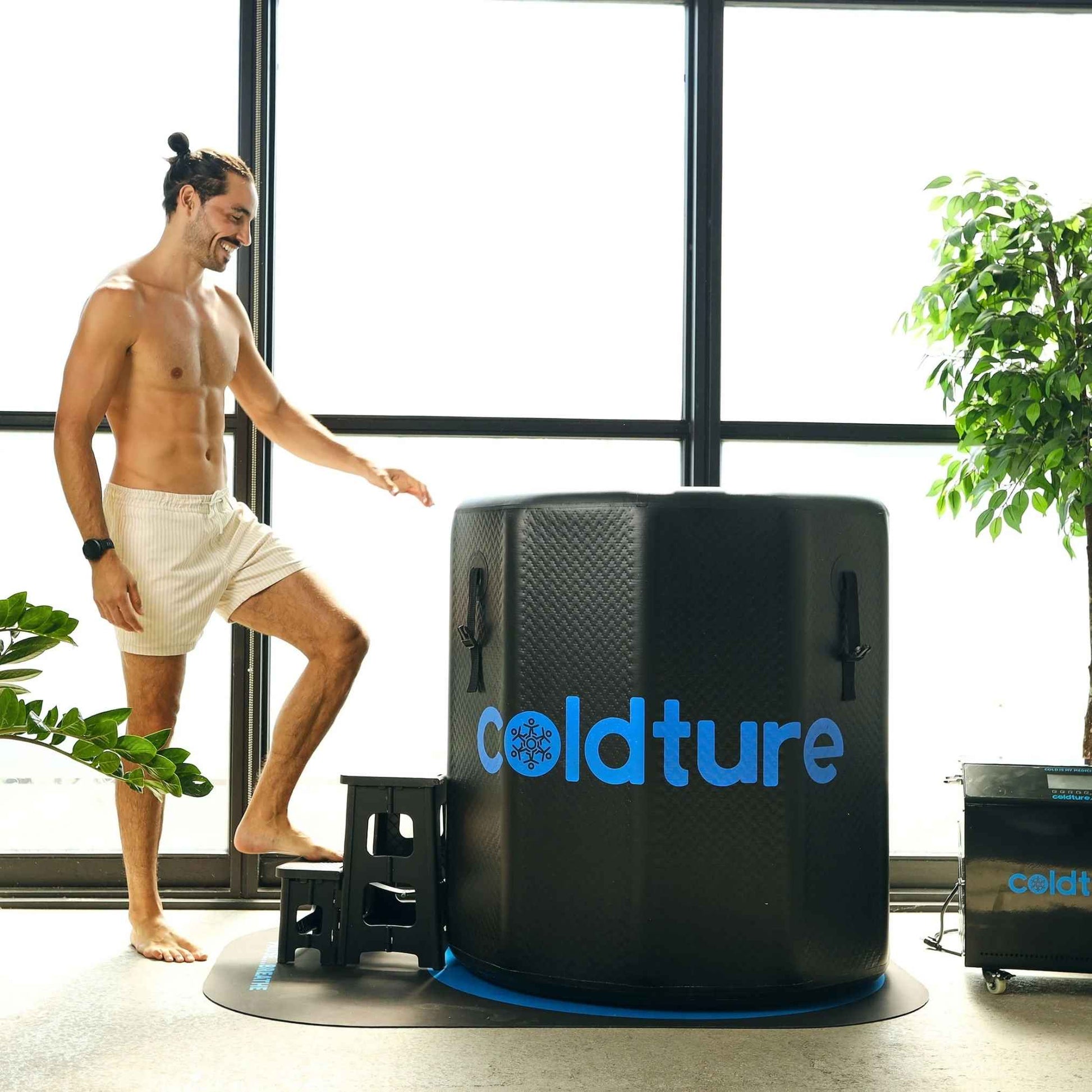 A man standing next to the Coldture Barrel + Chiller Pro, preparing to use the cold therapy system in a bright, indoor setting with plants.