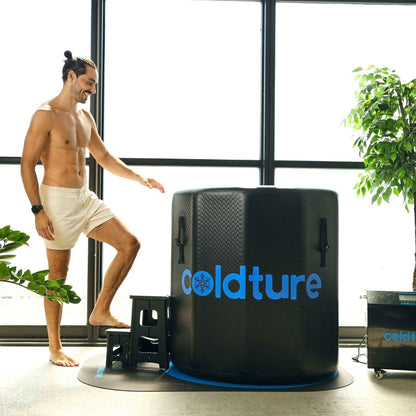 A man standing next to the Coldture Barrel + Chiller Pro, preparing to use the cold therapy system in a bright, indoor setting with plants.