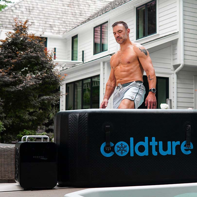 A shirtless man stepping into a Coldture Water Chiller Pro at a modern home, with a sleek design and a black exterior.