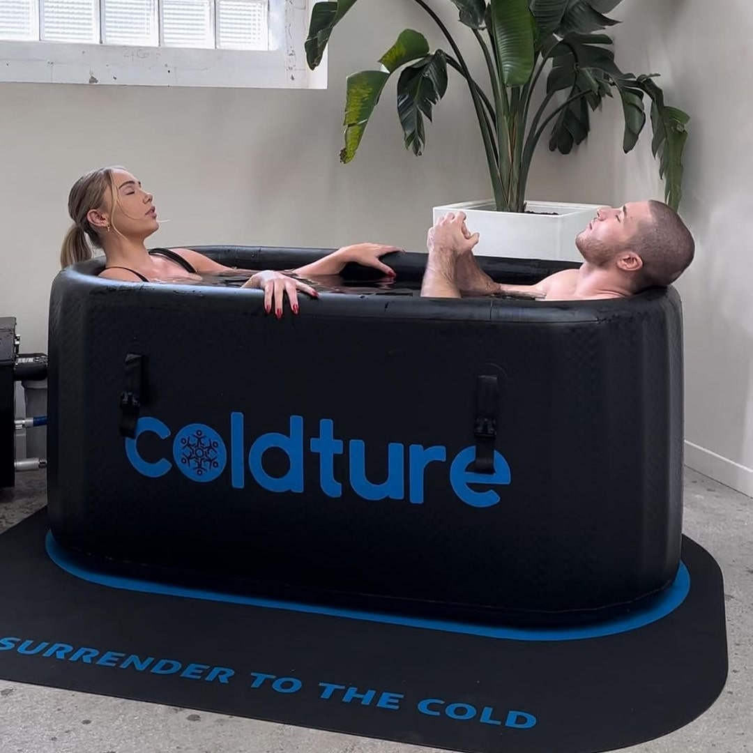 A woman and a man relaxing in a Coldture Classic Chiller Pro ice bath, with the text 'Surrender to the Cold' visible on the mat beneath them.