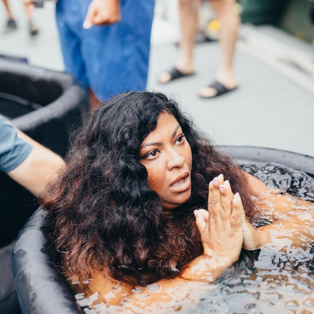 A woman with curly hair is sitting in a water-filled Coldture Classic + Chiller Pro tub, looking surprised or contemplative, with her hands together in a prayer-like gesture.