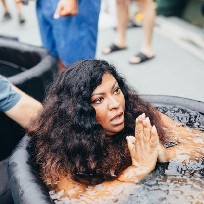 A woman with curly hair is sitting in a water-filled Coldture Classic + Chiller Pro tub, looking surprised or contemplative, with her hands together in a prayer-like gesture.