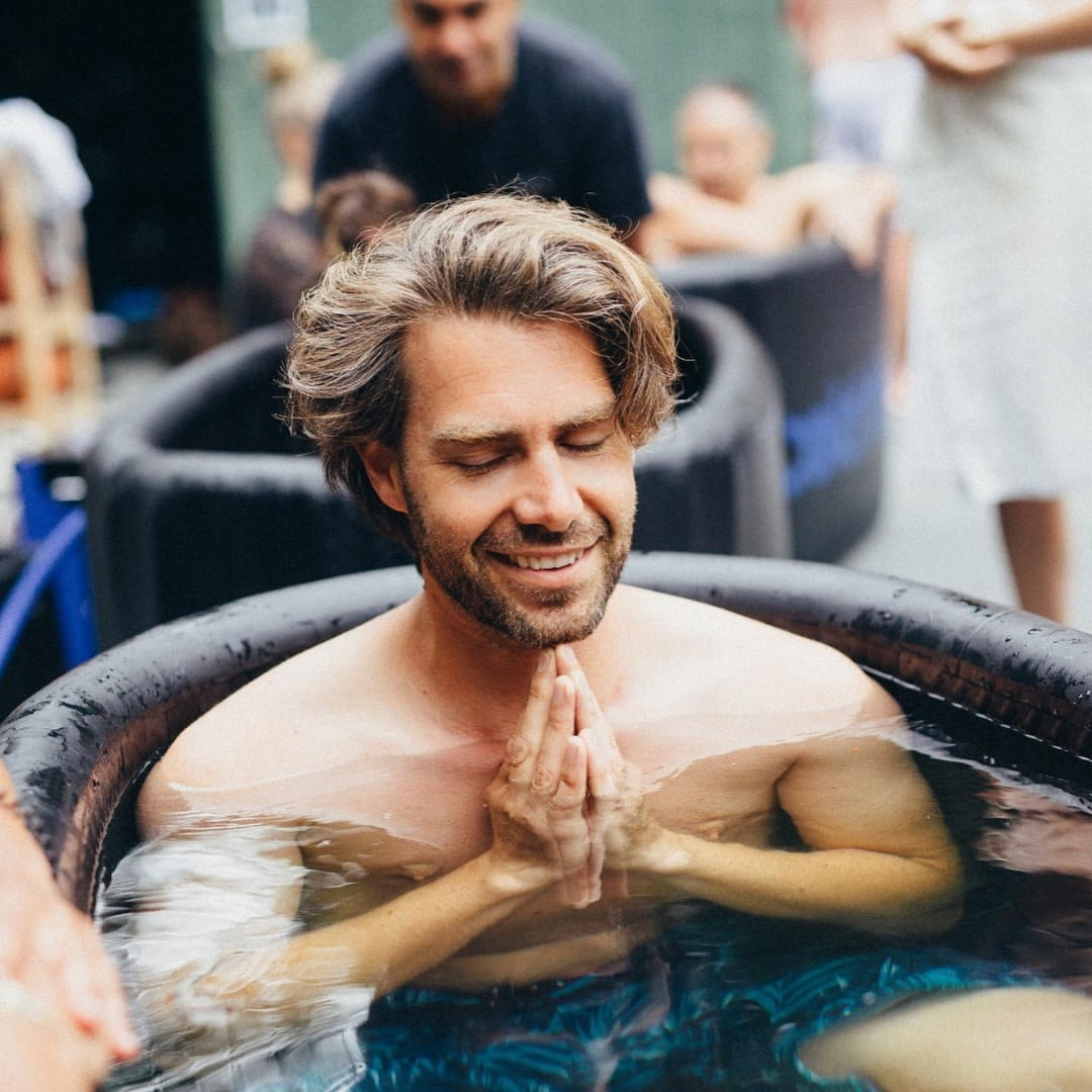 A man enjoying a moment of relaxation while seated in a Coldture Classic ice bath, featuring the Chiller Pro, with others in the background also participating in the experience.