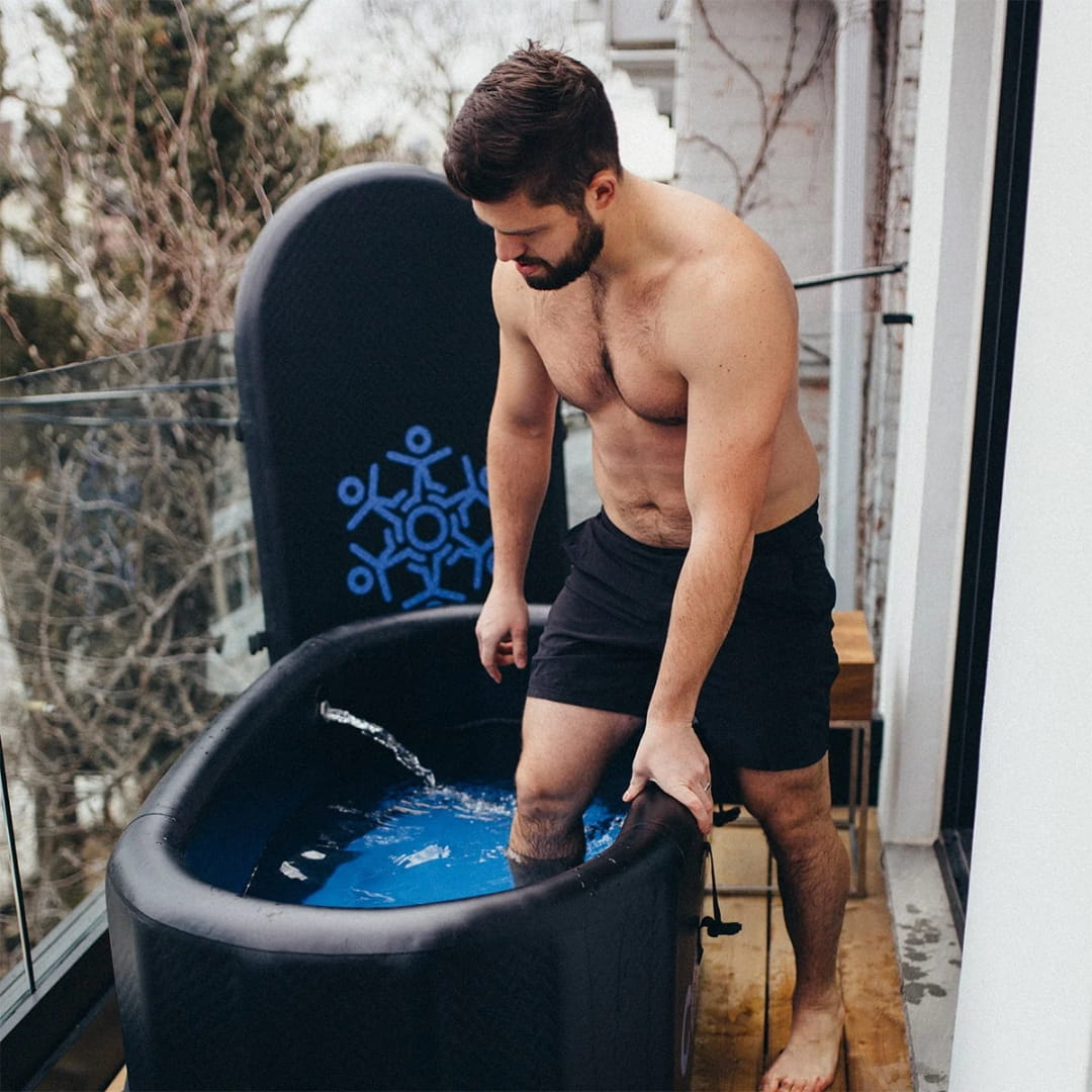 A man stands on a balcony, partially submerged in a Coldture Classic + Chiller Pro ice bath, with blue water and a modern design in the background.