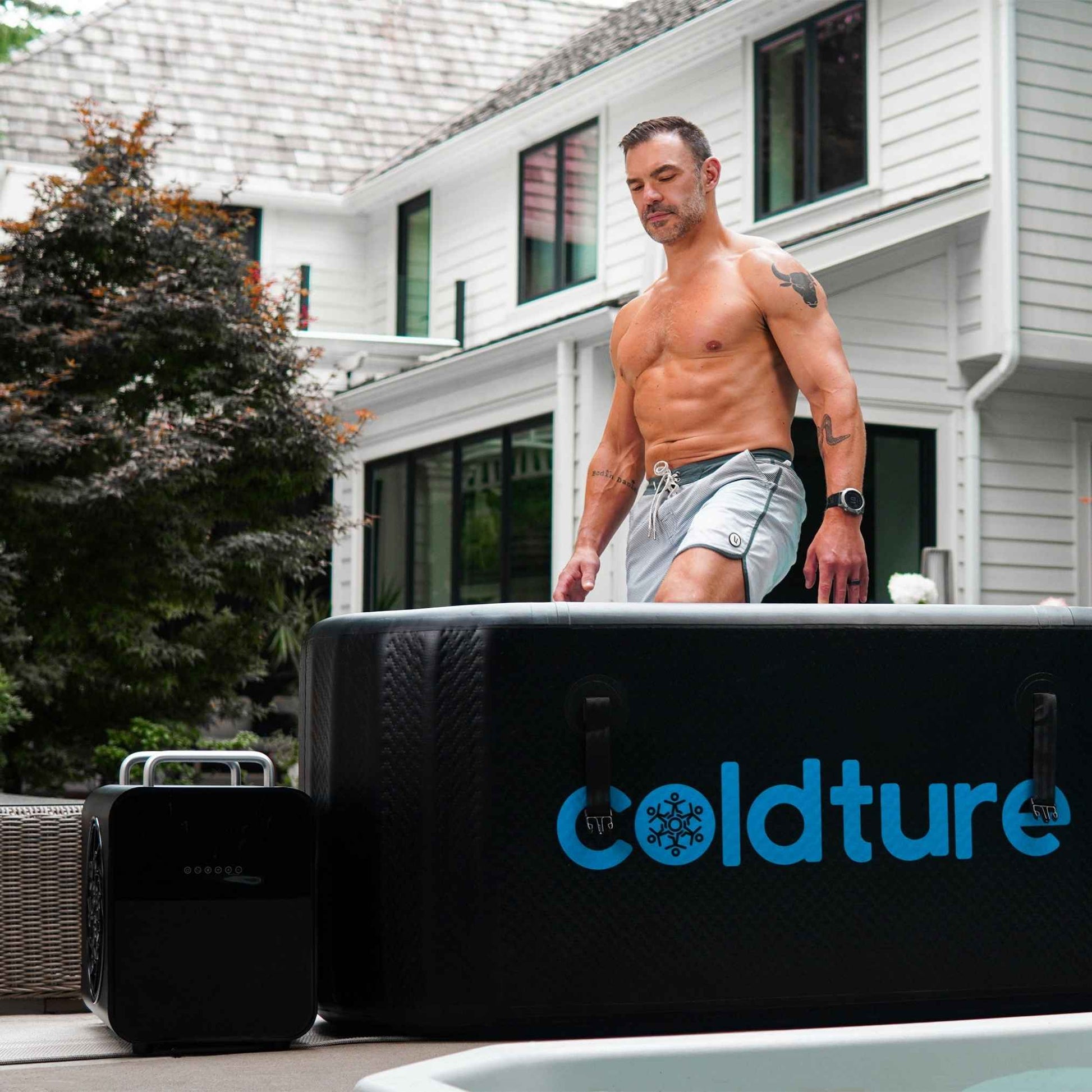 A man stepping into a Coldture Classic hot tub with a Chiller Pro unit beside it, set in a modern backyard.