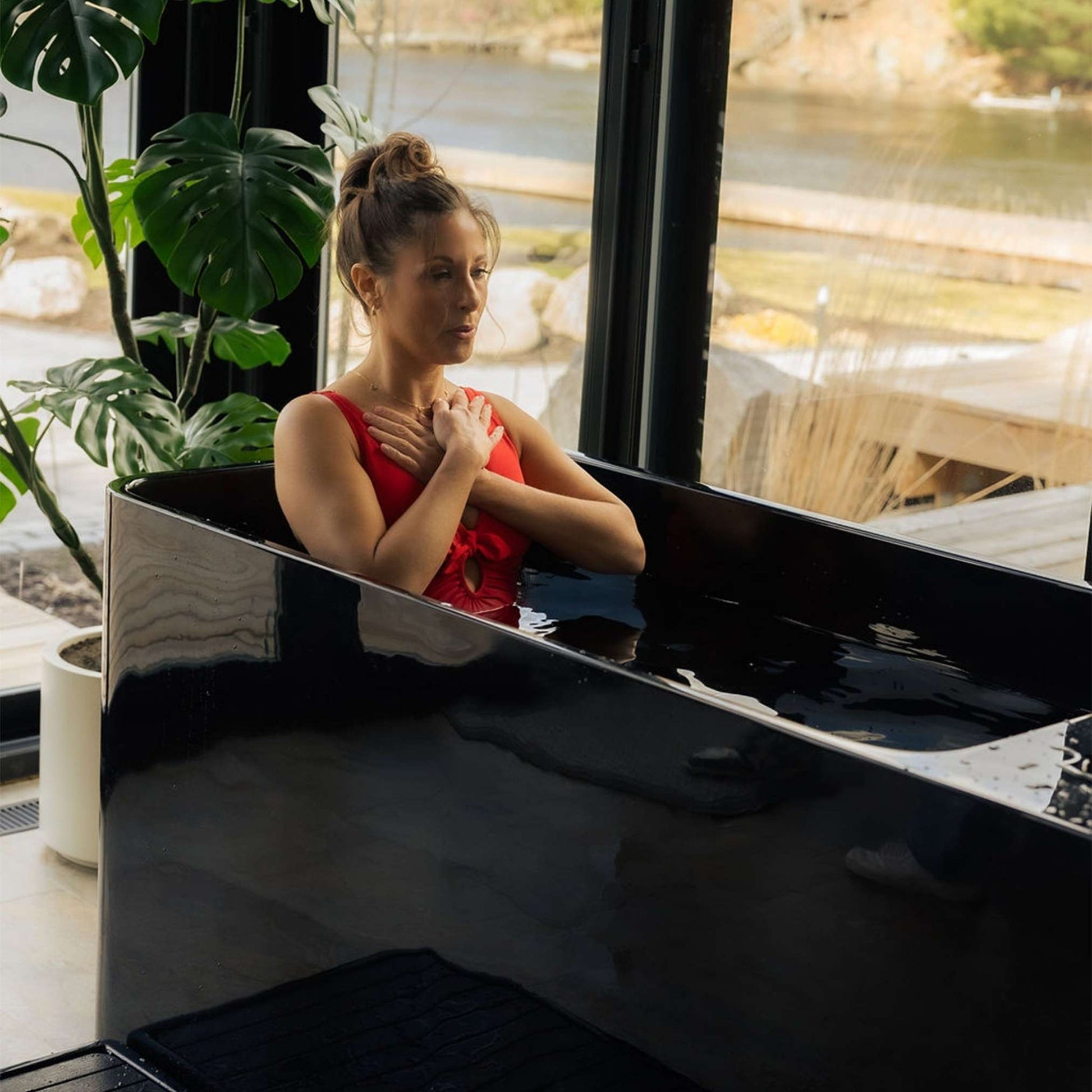 A woman in a red swimsuit is sitting in the Coldture Pro Plunge, immersed in water, with her hands on her chest, surrounded by large plants and a scenic view through the glass windows.