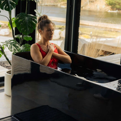 A woman in a red swimsuit is sitting in the Coldture Pro Plunge, immersed in water, with her hands on her chest, surrounded by large plants and a scenic view through the glass windows.