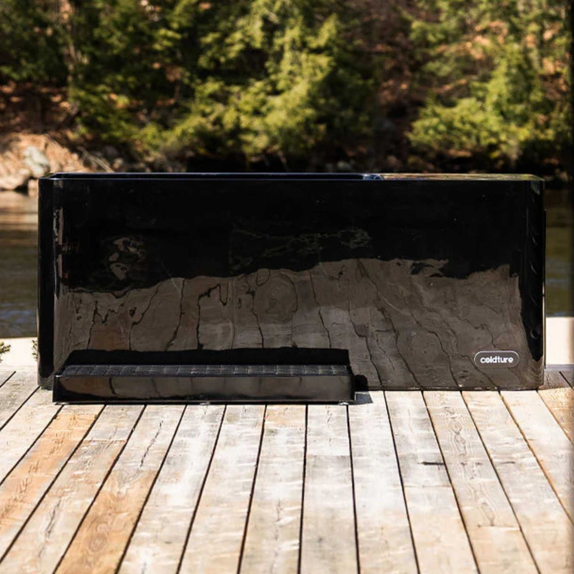 Coldture Pro Plunge, a sleek black plunge tub, positioned on a wooden deck by a tranquil water source, surrounded by lush greenery.