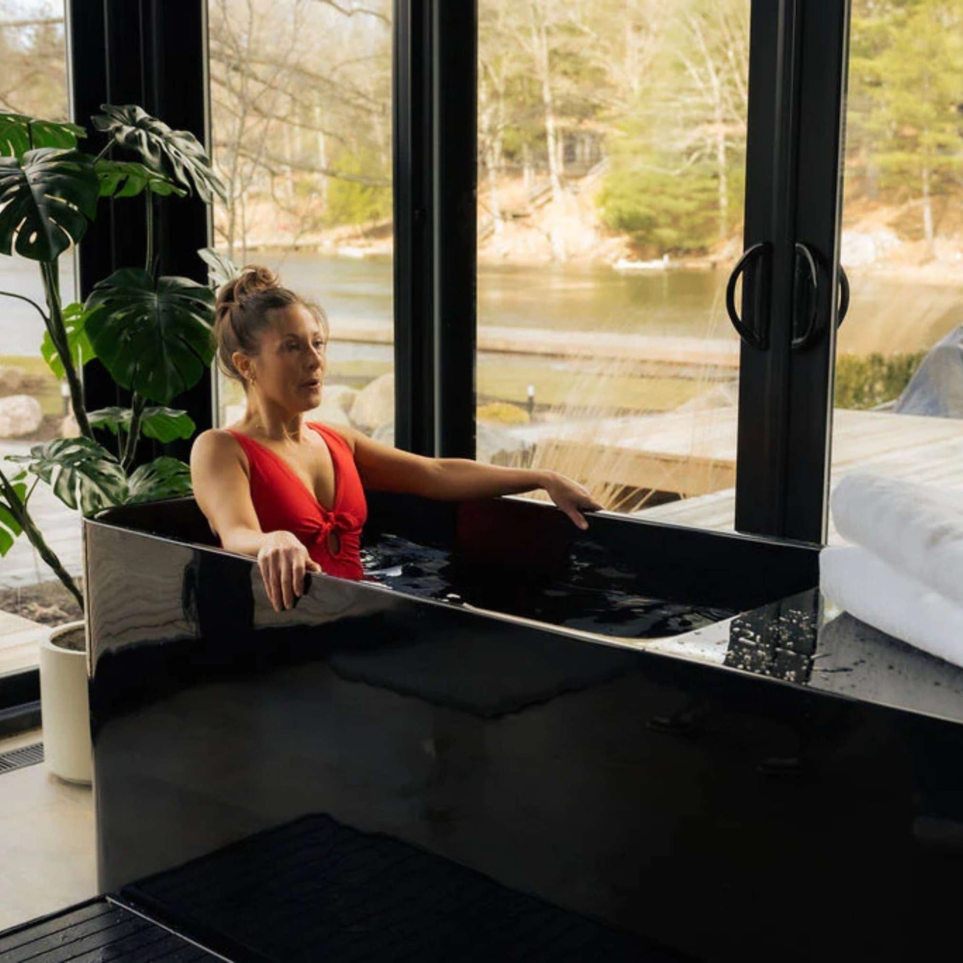 A woman in a red swimsuit relaxes in the Coldture Pro Plunge, an elegant black plunge tub, surrounded by large windows with a view of nature.