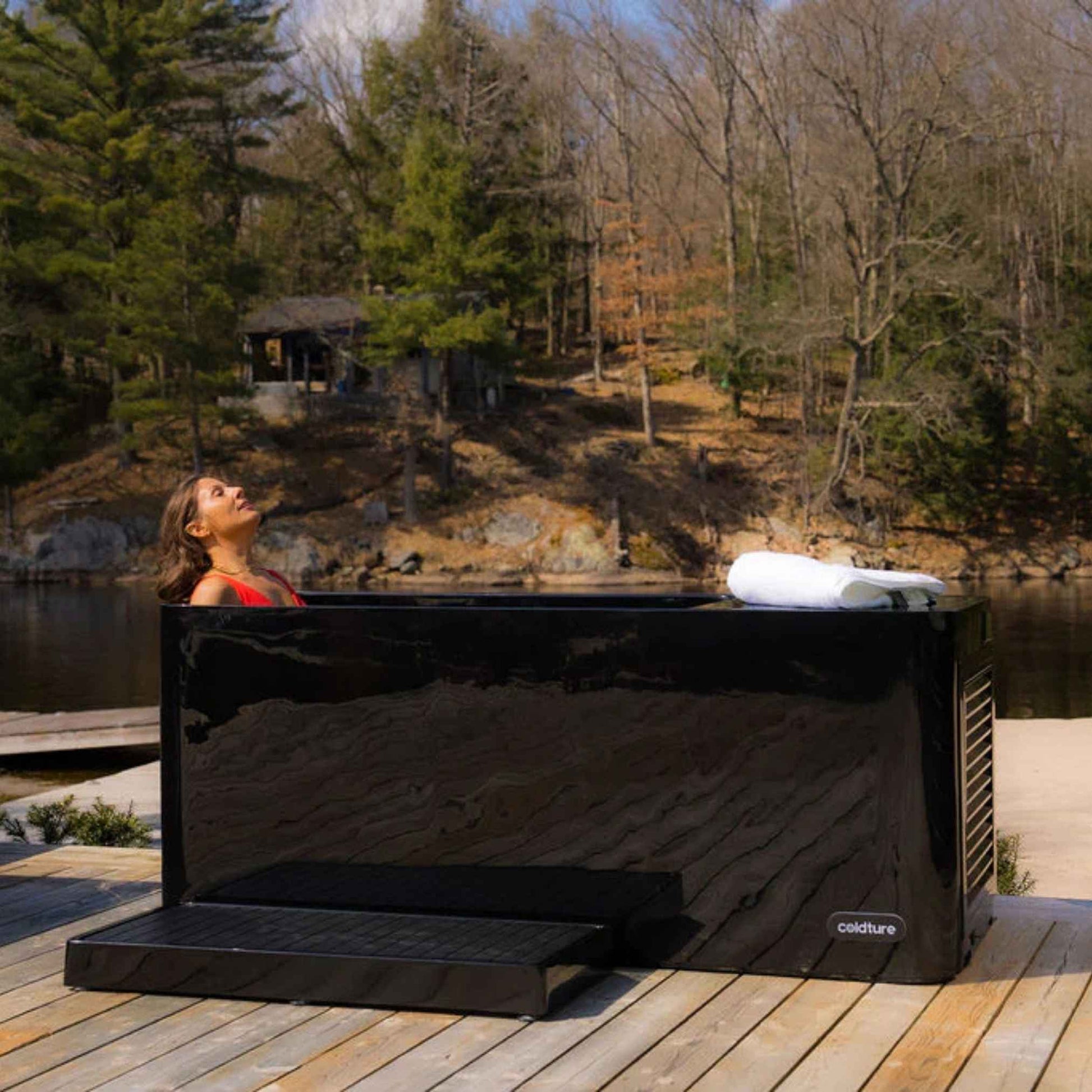 A woman relaxing in the Coldture Pro Plunge, a sleek black plunge tub, on a wooden deck by a serene lake surrounded by trees. A white towel rests on the edge of the tub, adding to the tranquil atmosphere.