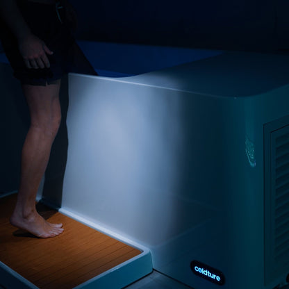 A person standing beside the Coldture Pro Plunge, with one foot on the wooden platform and the other poised to enter, illuminated by soft lighting in a dark environment.
