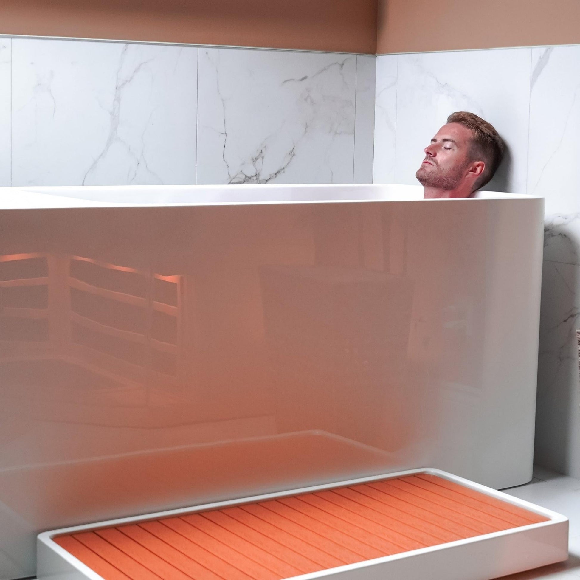 A person relaxing in a Coldture Pro Plunge bath, with a modern design and marble wall backdrop.