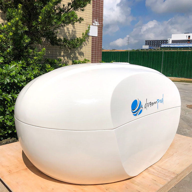 Dreampod Home Float Plus, a sleek white flotation pod, positioned outdoors with greenery in the background.